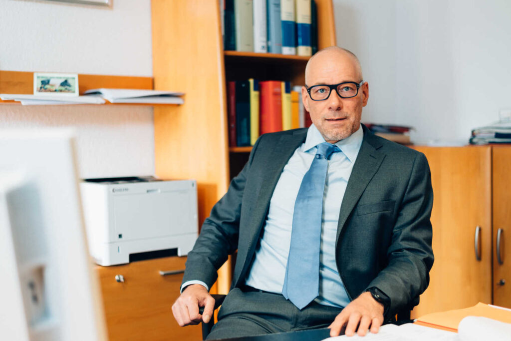 Ein Mann mit kurzen Haaren, einer dunklen Brille und einem dunklen Anzug sitzt an seinem Schreibtisch im Büro.